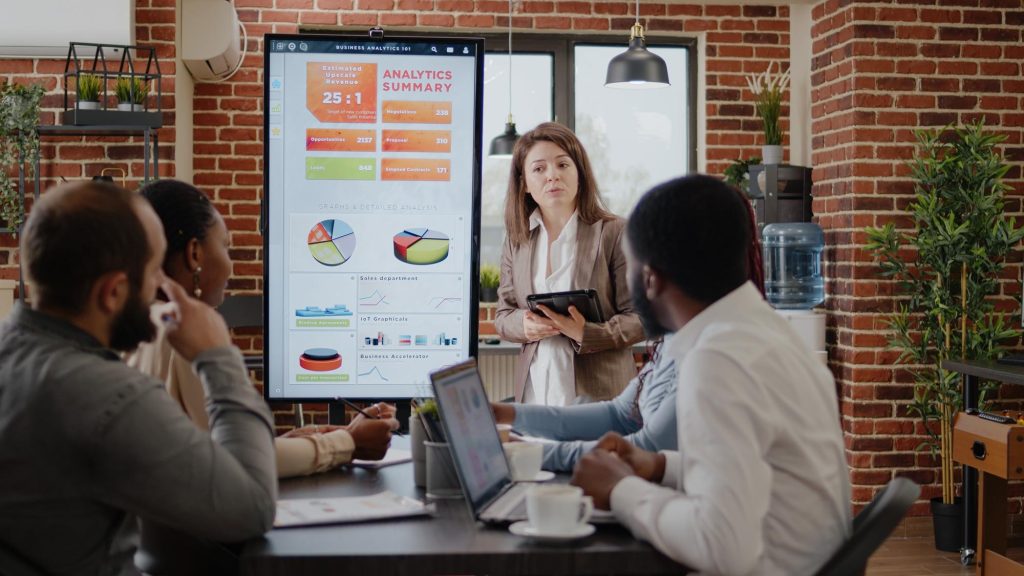 Business meeting with woman presenting analytics summary on screen, discussing AEO trends and SEO strategies with attentive colleagues.